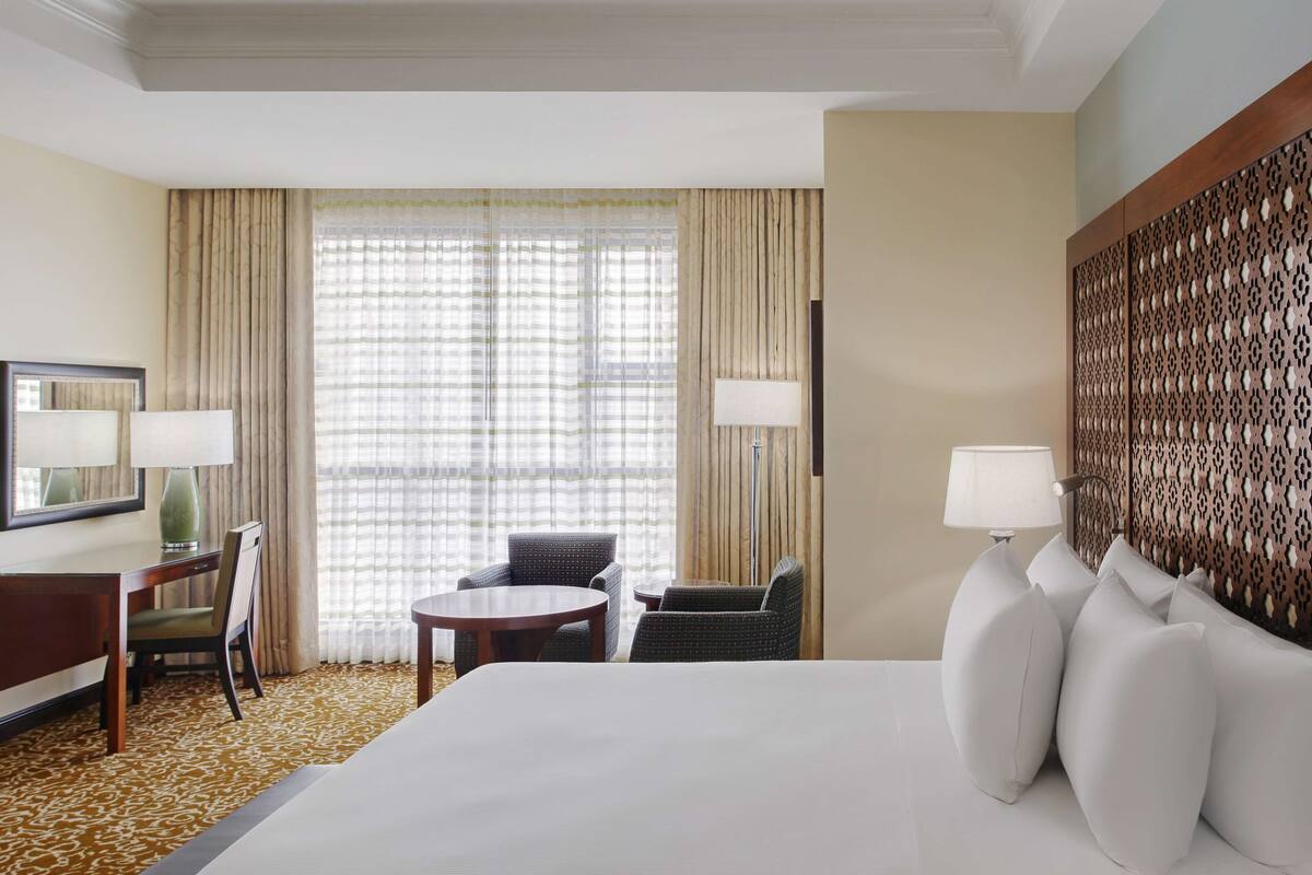 Habitación de hotel con una cama grande, dos sillas, una mesita, un escritorio, una lámpara y un ventanal con cortinas.