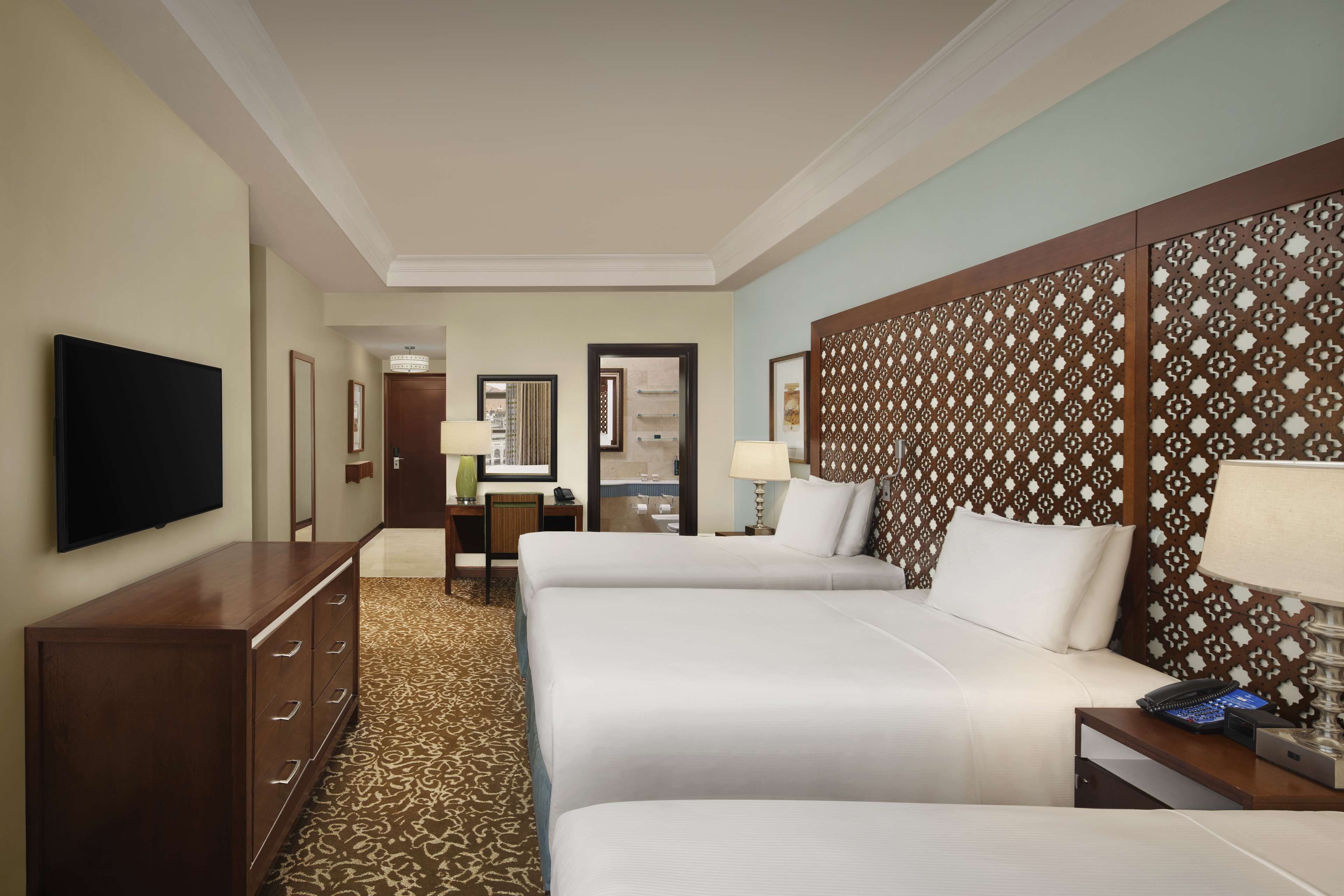 A hotel room with two beds, a TV, a desk, and a patterned headboard.