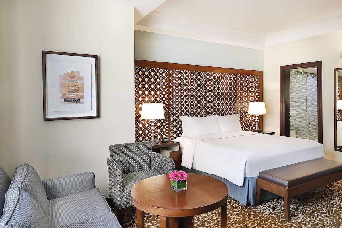 Habitación de hotel con cama, dos mesitas de noche, una silla, una mesa redonda con un florero y un cuadro en la pared.