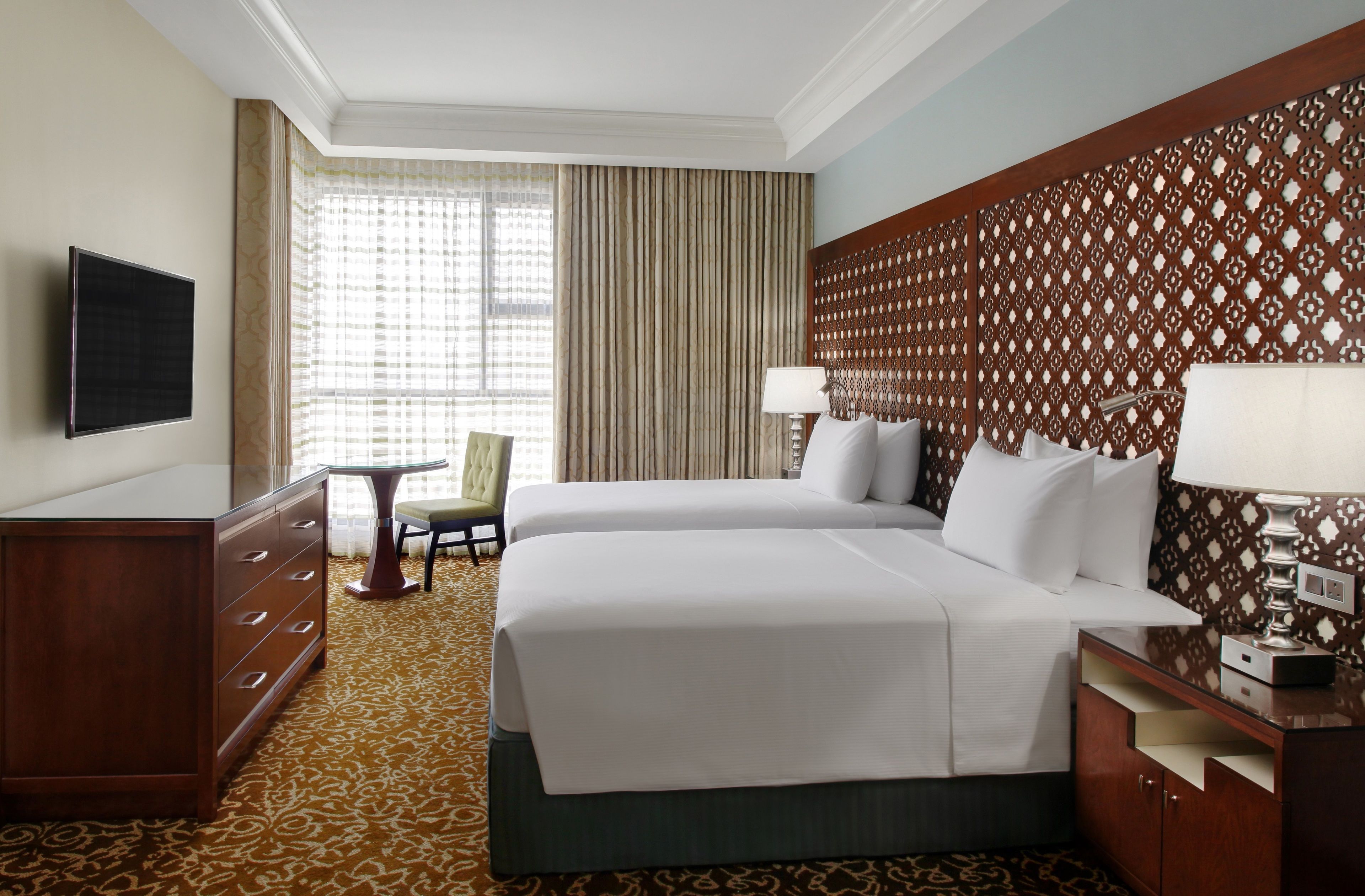 A hotel room with two beds, a TV, a desk, a chair, and a patterned wall.