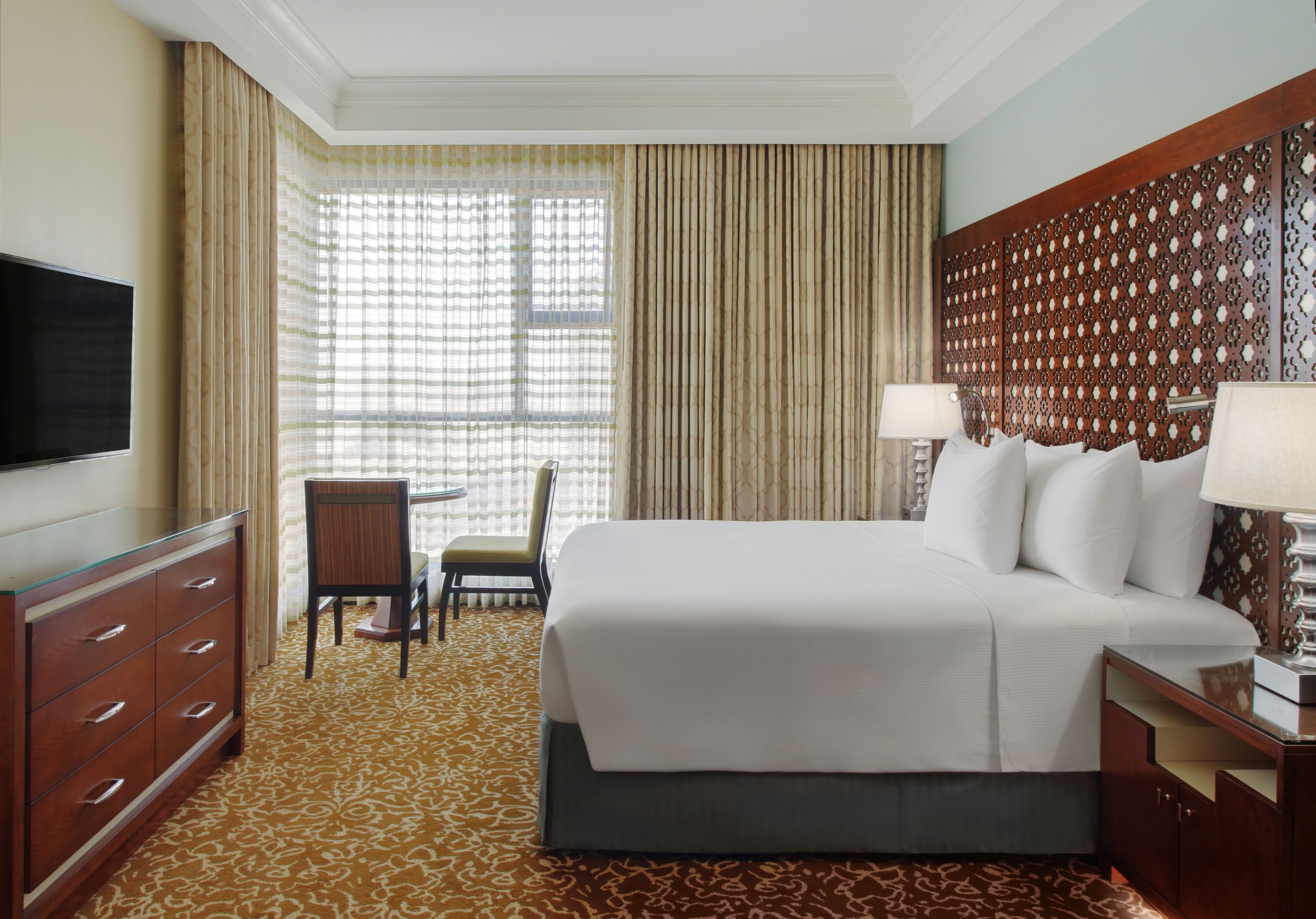 Quarto de hotel com uma cama grande, televisão, escrivaninha com cadeira e tapete estampado.
