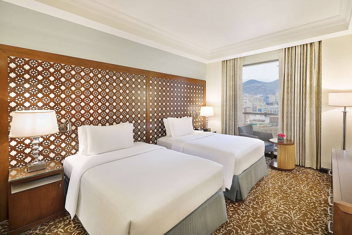 Habitación de hotel con dos camas, un cabecero de madera decorativo, vistas a la ciudad y una alfombra estampada.