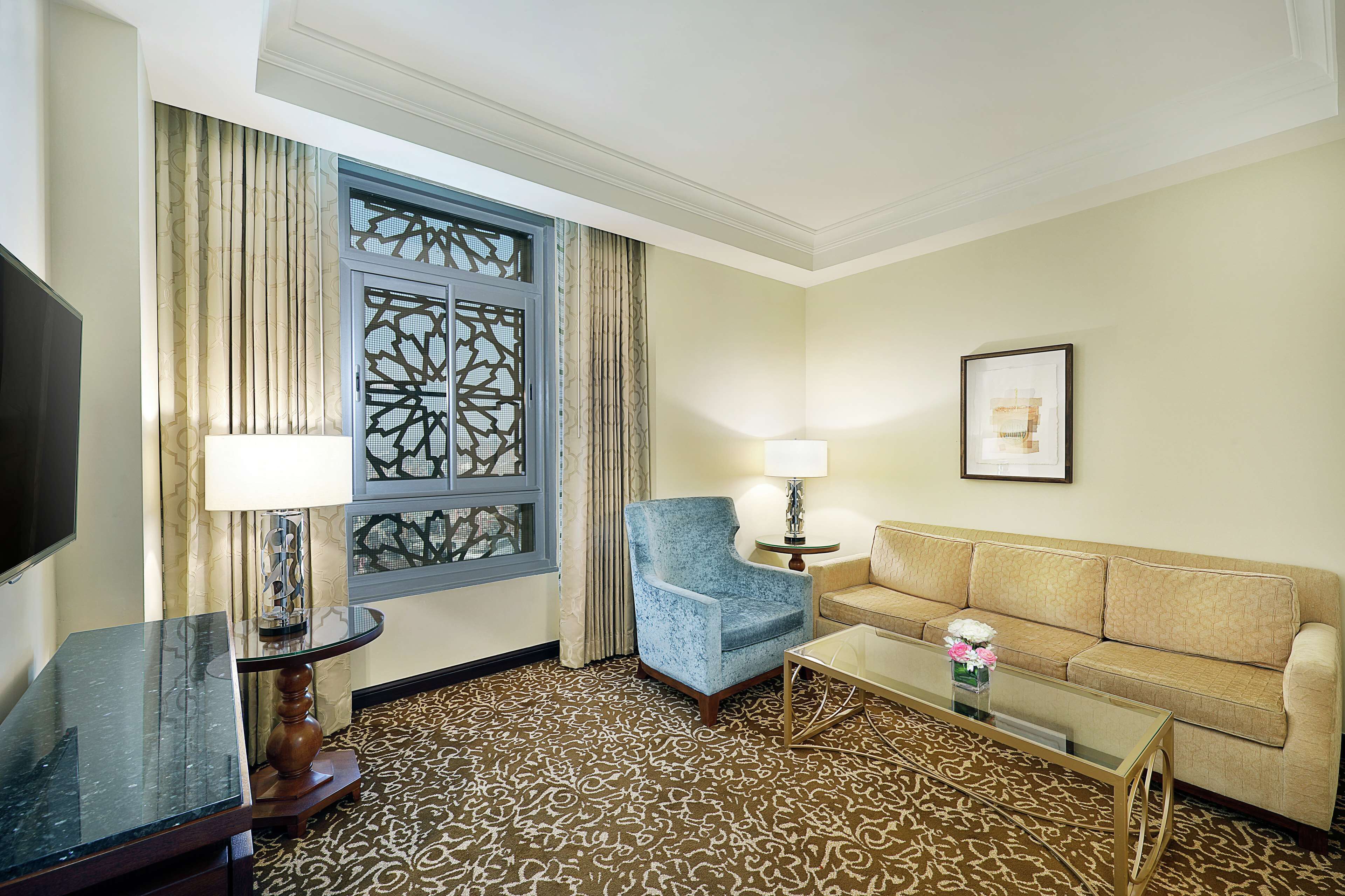 A hotel room with a patterned carpet, a blue armchair, a beige sofa, a glass coffee table, a TV, and a decorative window with curtains.