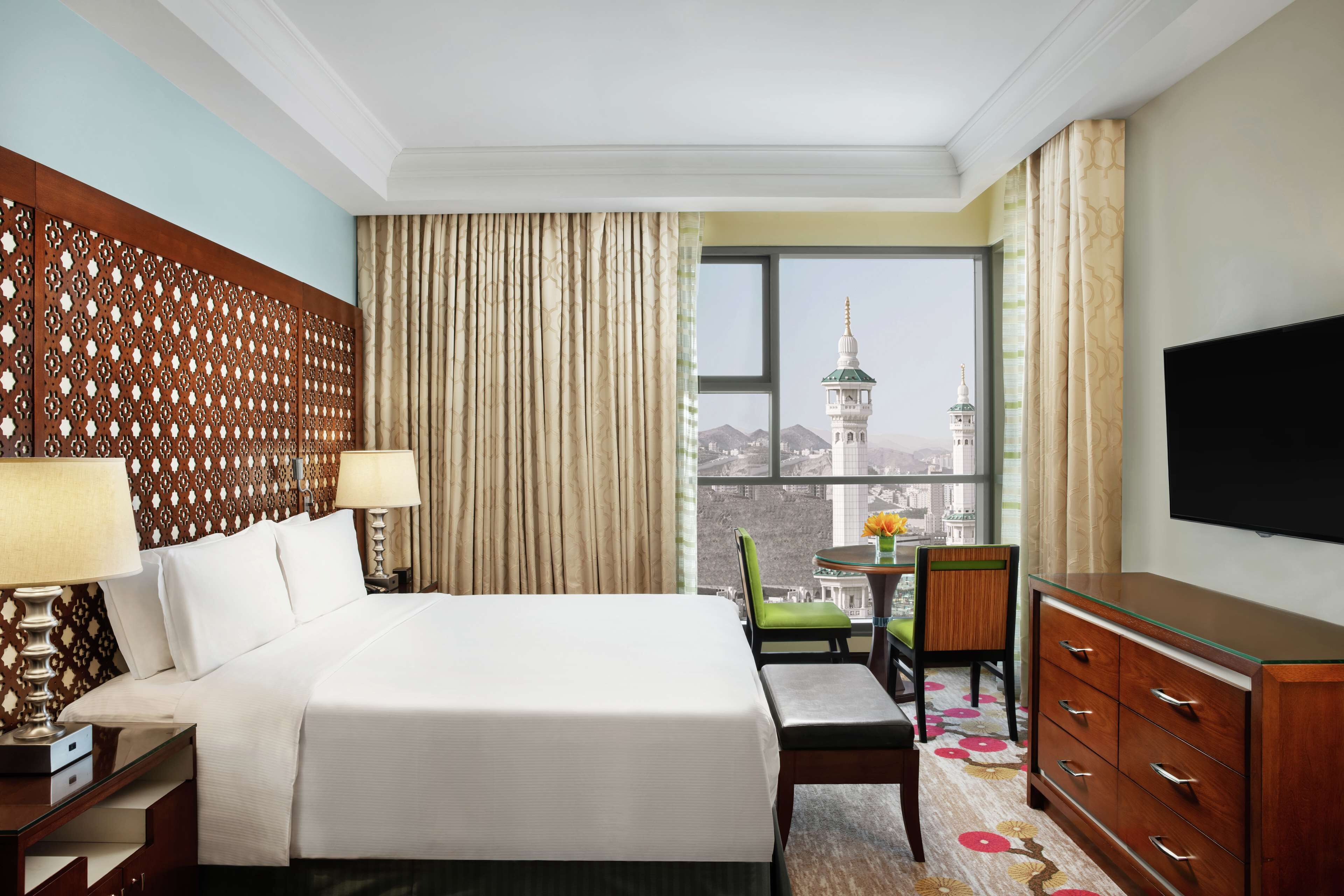 A hotel room with a large bed, a desk with a chair, a television, and a view of a mosque with a minaret.