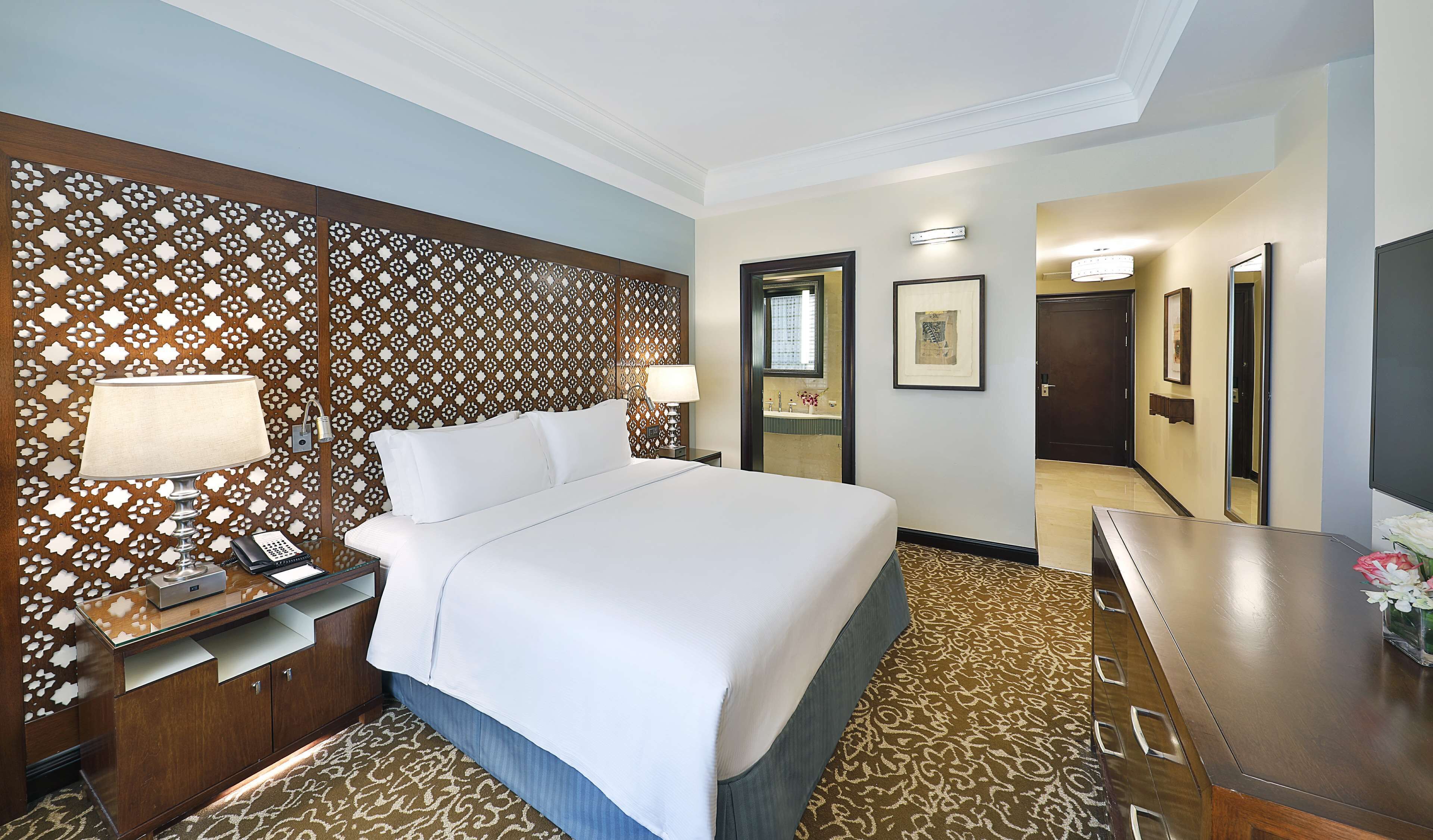 A hotel room with a large bed, a wooden headboard with a patterned design, a bedside table with a lamp, a phone, and a small cabinet. There is a mirror on the wall, a door leading to another room, and a carpeted floor.