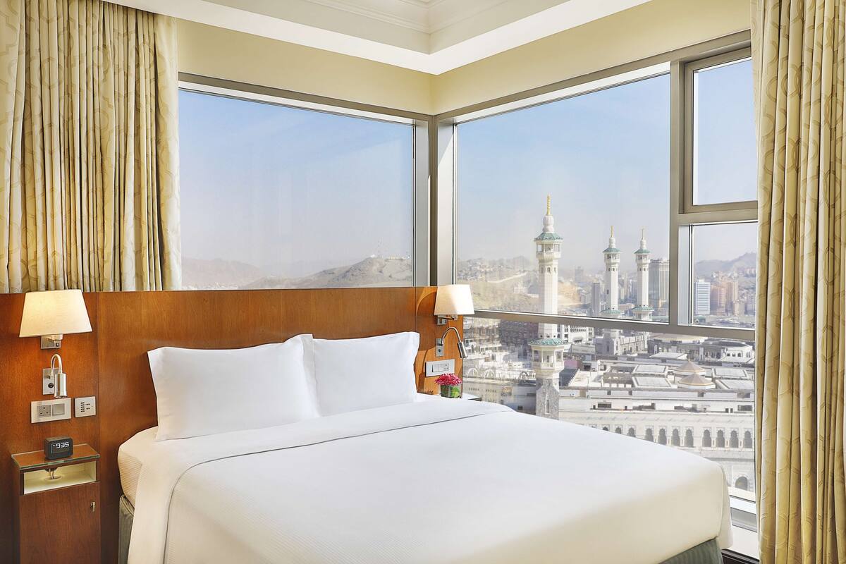Habitación de hotel con una cama grande, una mesita de noche con lámpara y vistas a un paisaje urbano con una torre.
