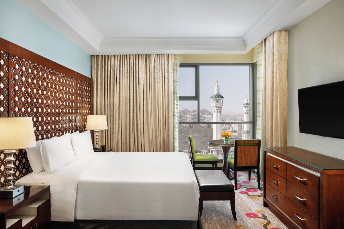 A hotel room with a large bed, a desk with a lamp, a dresser, and a television. There is a window with a view of a cityscape and a mosque.