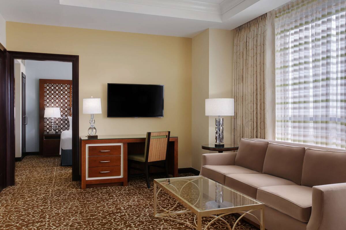 Habitación de hotel con sofá, escritorio y silla, televisor y ventana con cortinas.