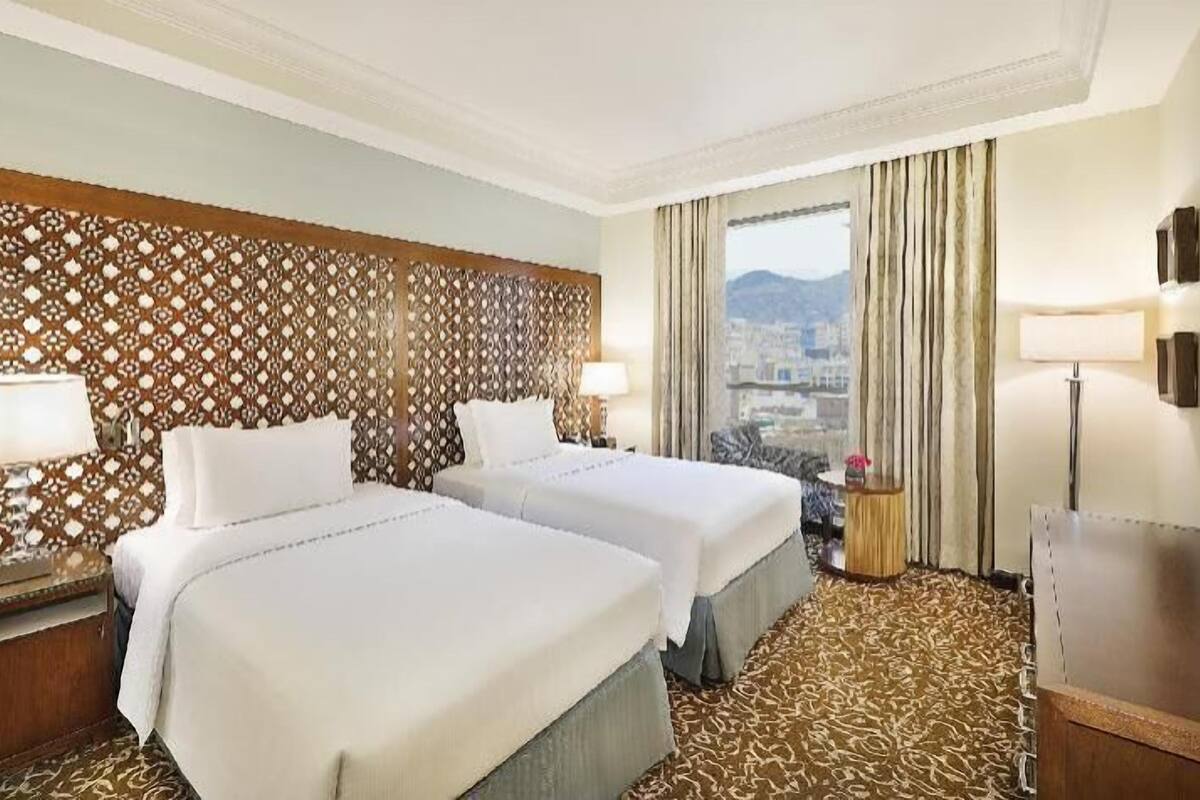 Habitación de hotel con dos camas, un cabecero estampado, vistas a la ciudad y un escritorio con lámpara.