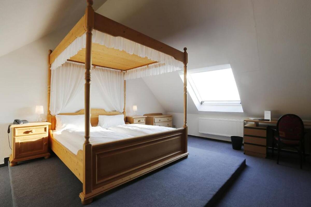 Een hemelbed met witte lakens, een houten hoofdbord en een nachtkastje met een lamp. Er is een bureau met een stoel en een radiator in de hoek.