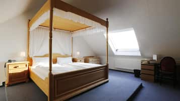 A four-poster bed with white linens, a wooden headboard, and a nightstand with a lamp. There is a desk with a chair and a radiator in the corner.