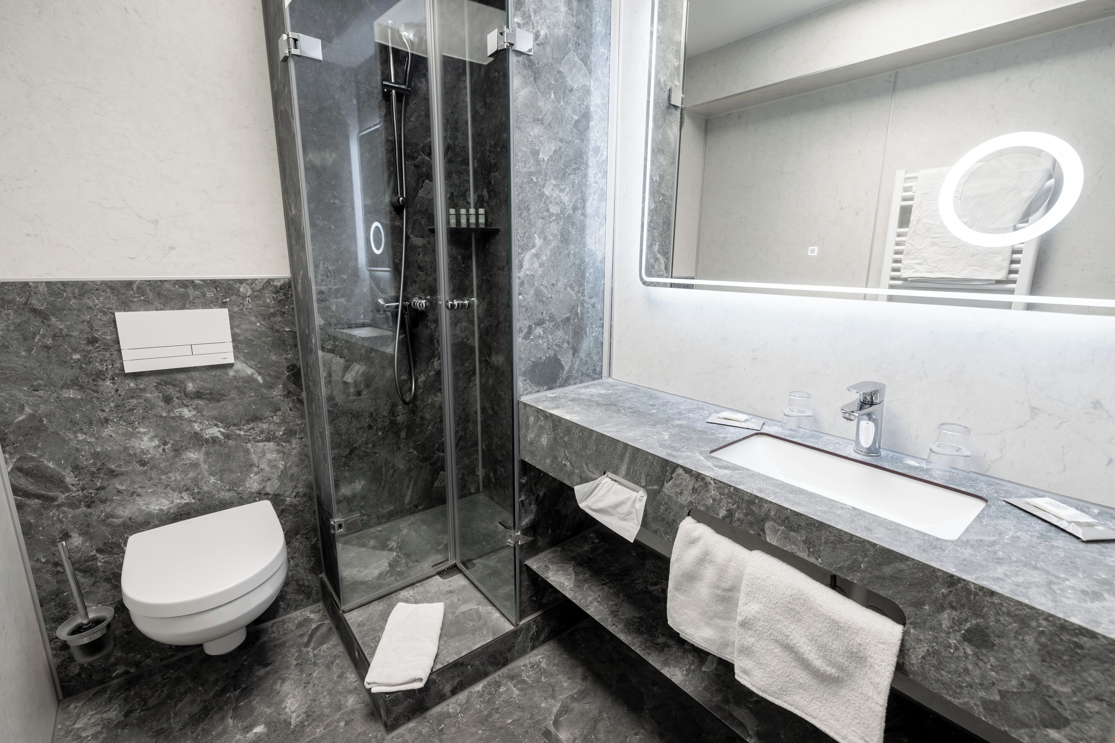 Ein modernes Badezimmer mit einer gläsernen Duschkabine, einer weißen Toilette, einer Marmorarbeitsplatte und einem großen Spiegel mit runder Beleuchtung.