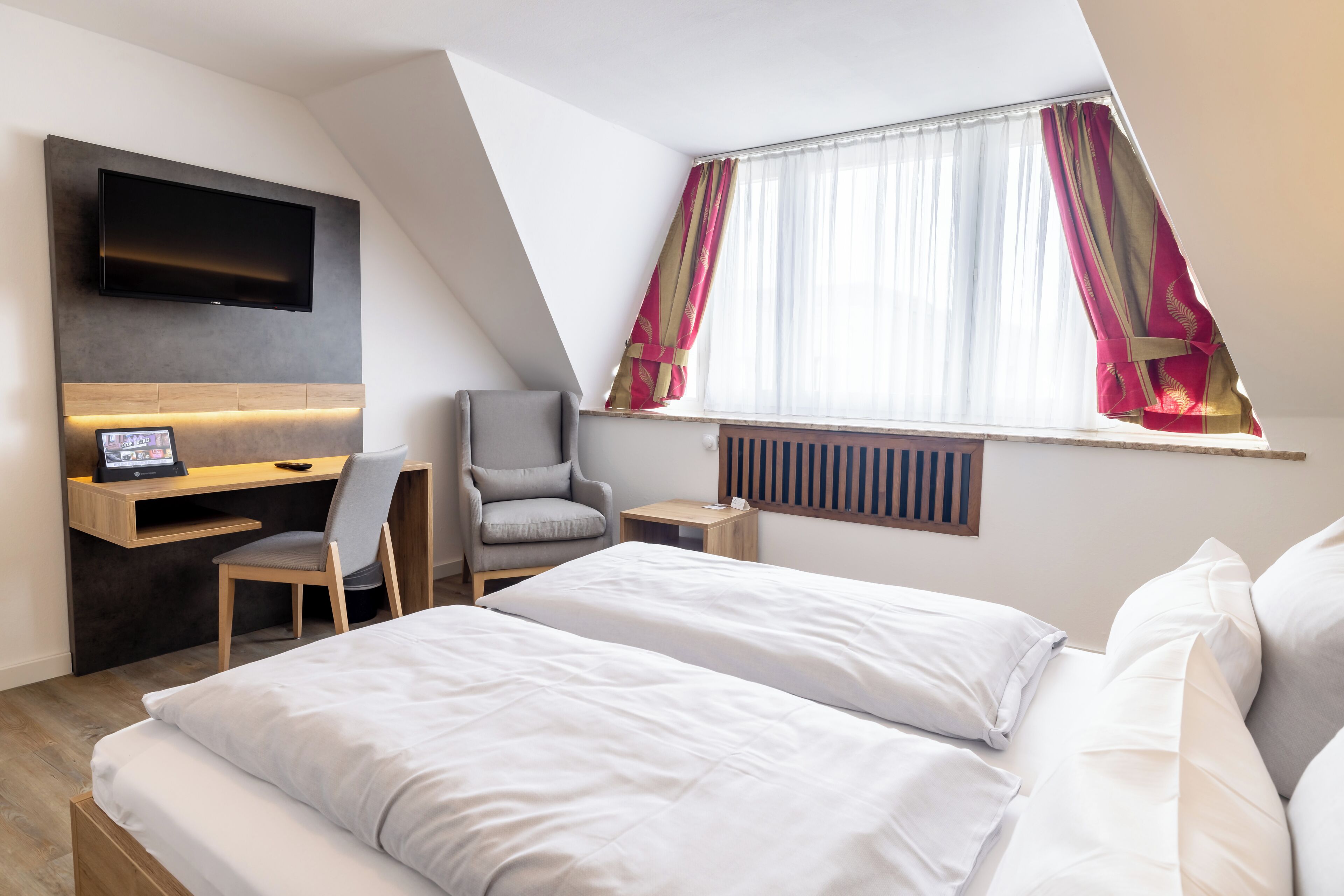 Ein Hotelzimmer mit Bett, Schreibtisch mit Computer, Stuhl, Wandfernseher und einem fensterbepflanzten Fenster.