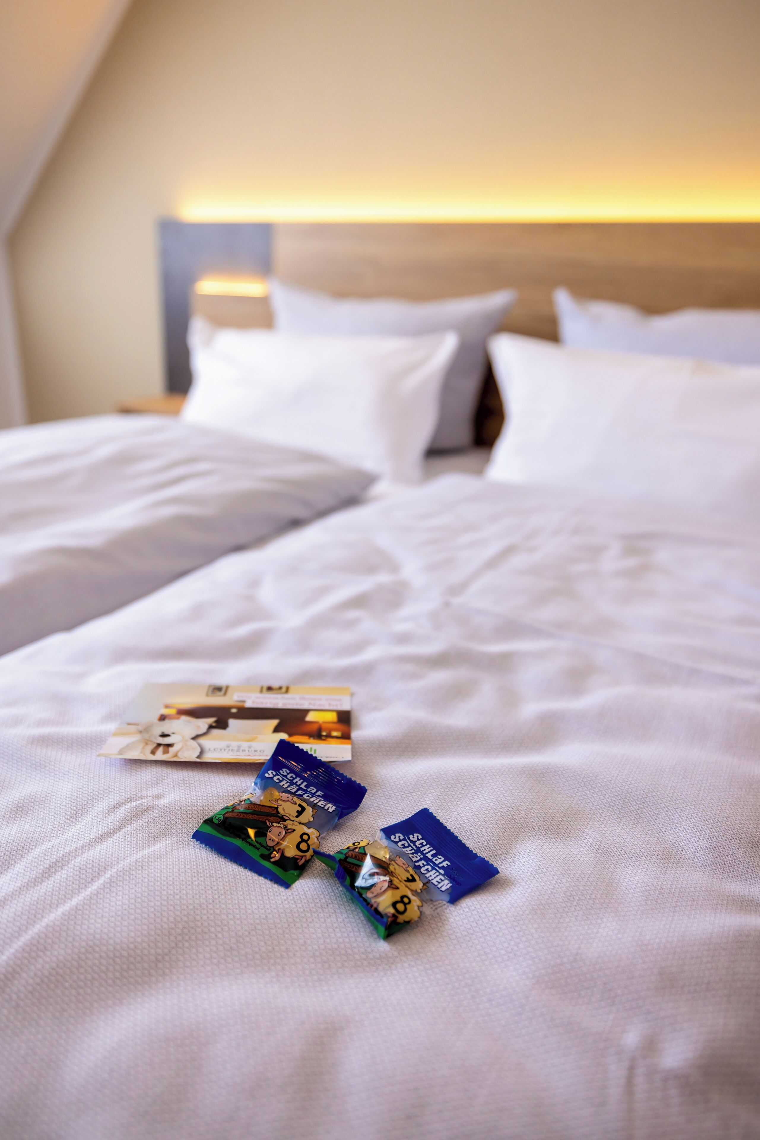 A neatly made bed with white linens and pillows, two travel brochures on the bed, and a headboard with a light fixture.