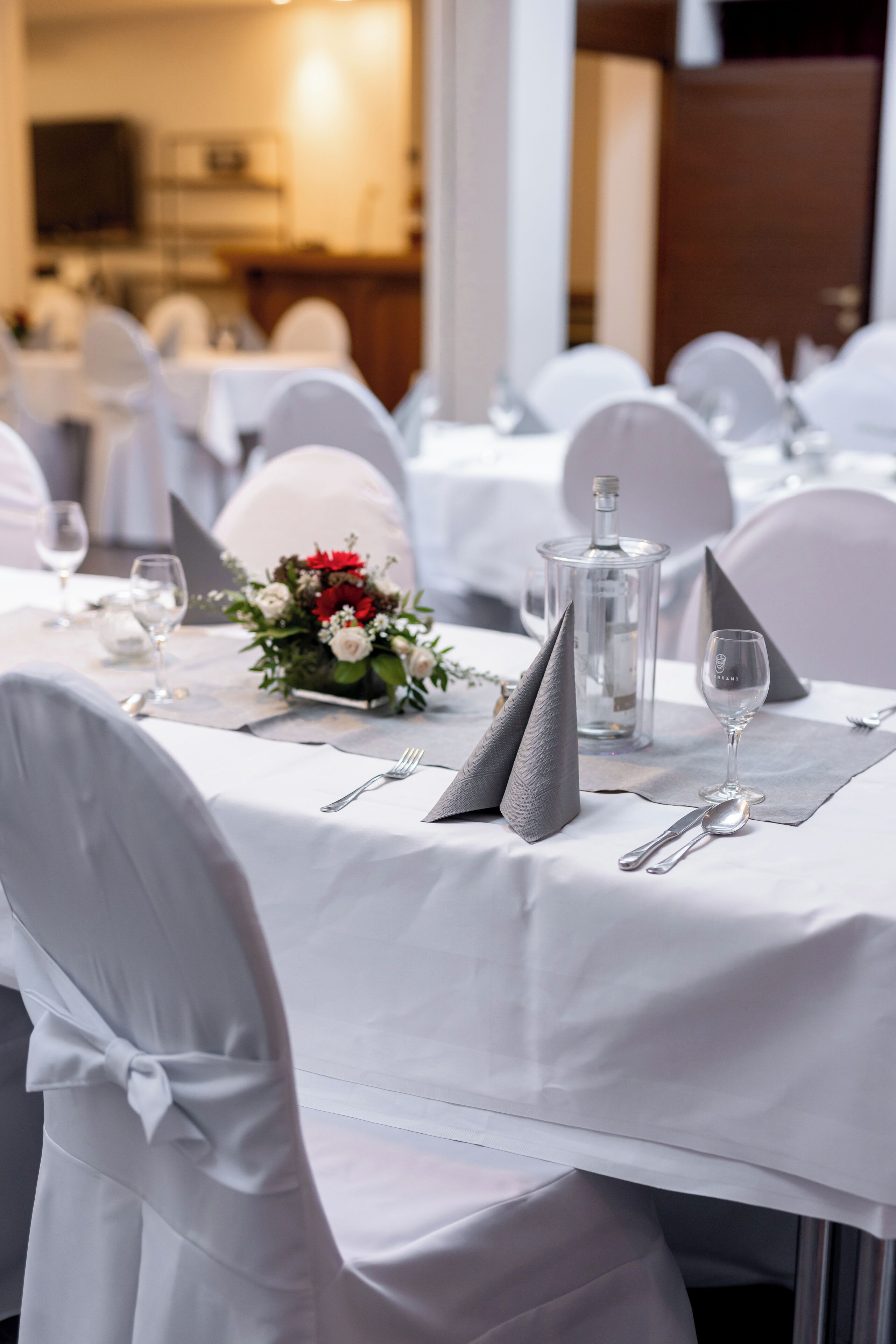 A dining area with white tablecloths, neatly set tables with silverware, glasses, and a centerpiece with flowers.