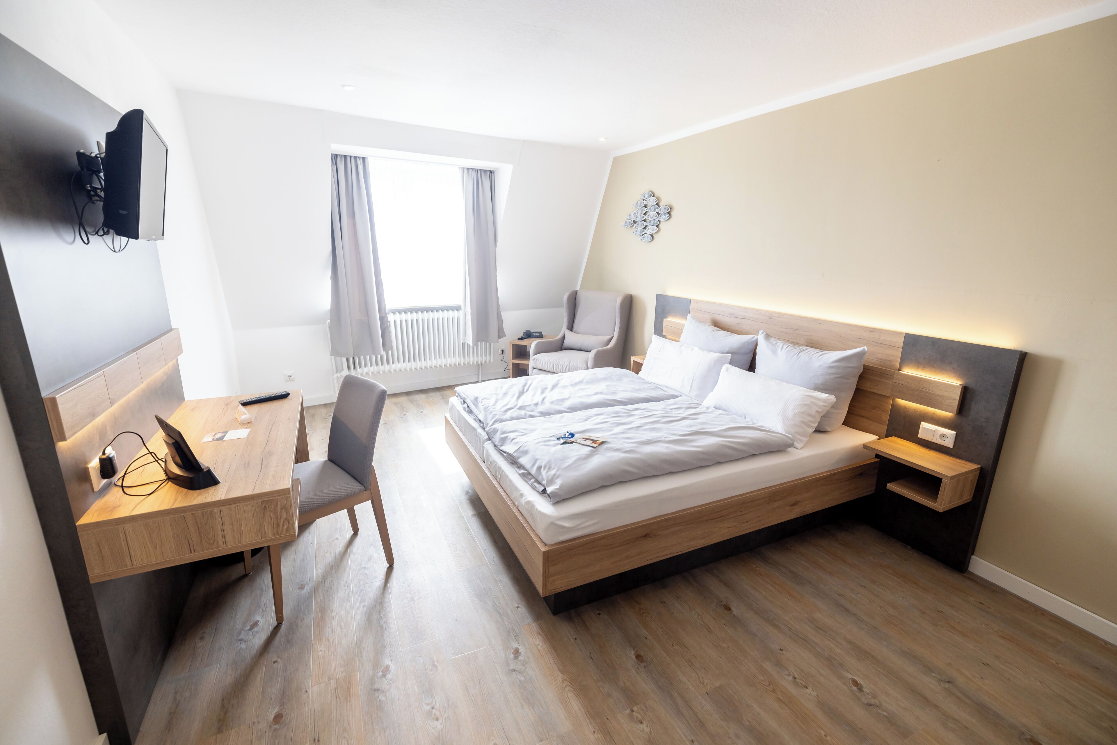 Quarto de hotel moderno com cama de madeira, mesas de cabeceira, escrivaninha com cadeira, televisão na parede e janela com cortinas.