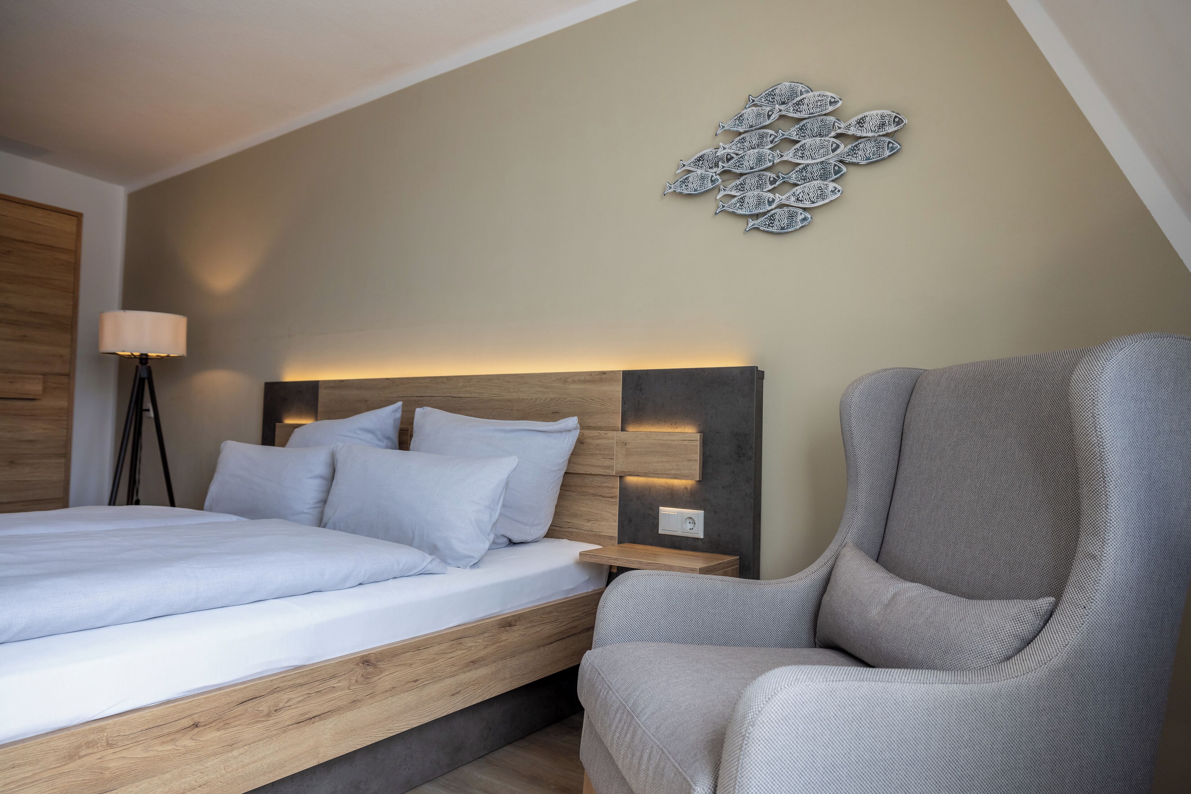 A modern bedroom with a wooden headboard, a grey upholstered armchair, and a wall-mounted decorative fish art piece.