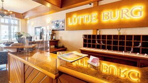 Un espace d’accueil avec un comptoir en bois, une grande enseigne lumineuse portant l’inscription « LUTTJE BURG » et une vitrine présentant des verres à vin.