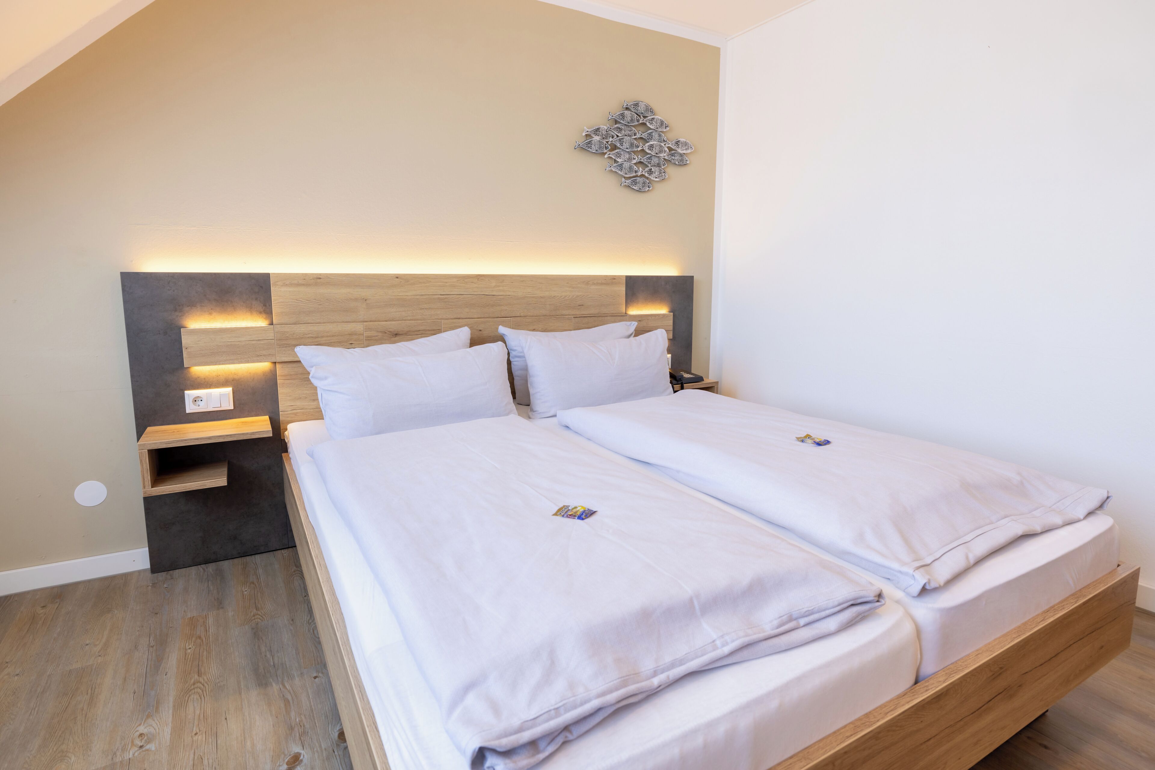 Una cama doble con cabecera de madera y mesitas de noche, un adorno en la pared, y un suelo de tono claro.