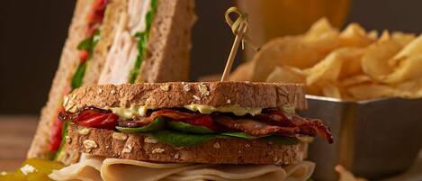 A sandwich with a toothpick, a glass of beer, and a bowl of chips on a wooden table.