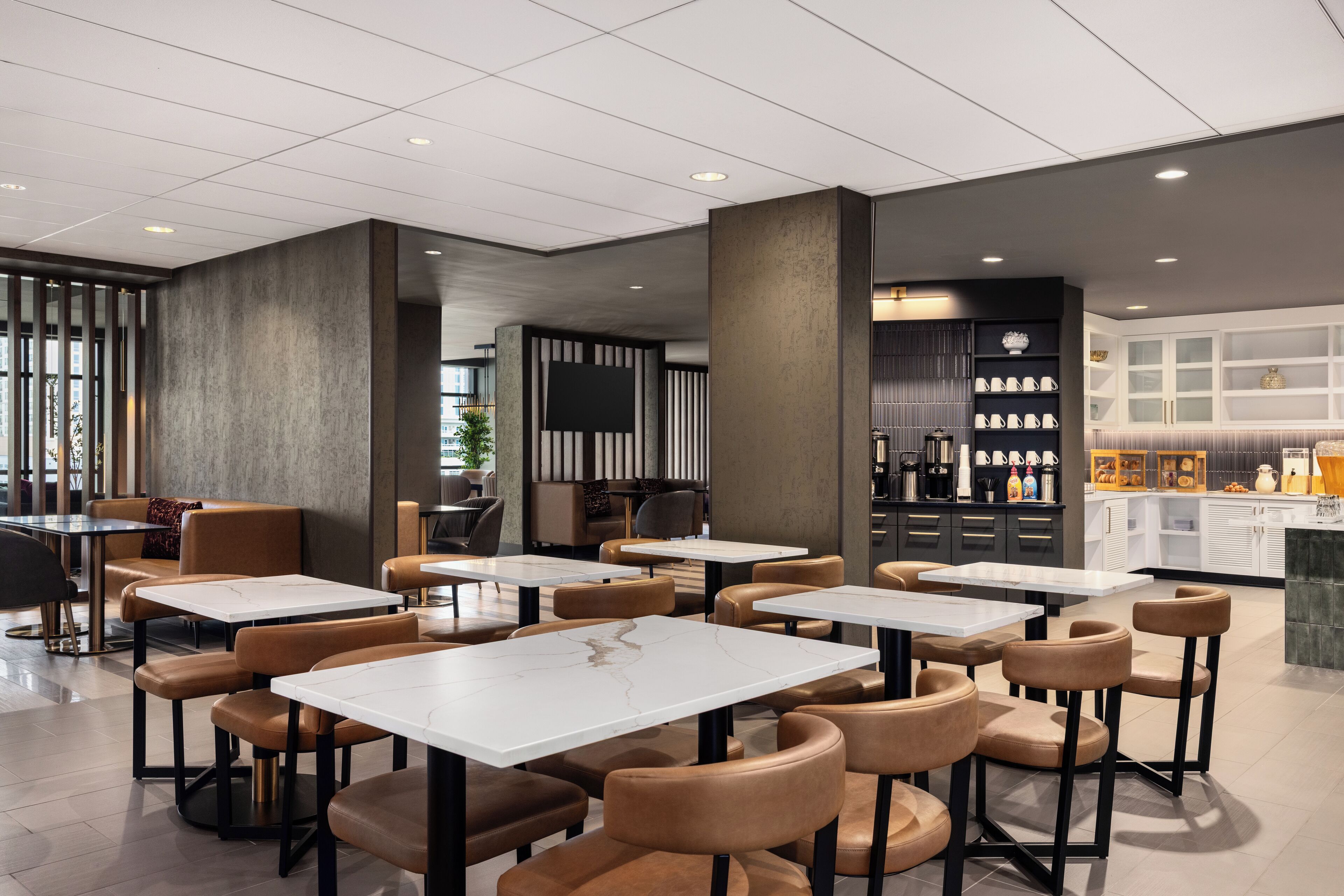 A modern cafe with white marble tables and brown leather chairs, featuring a kitchen area with shelves of dishes and a seating area with a large window.