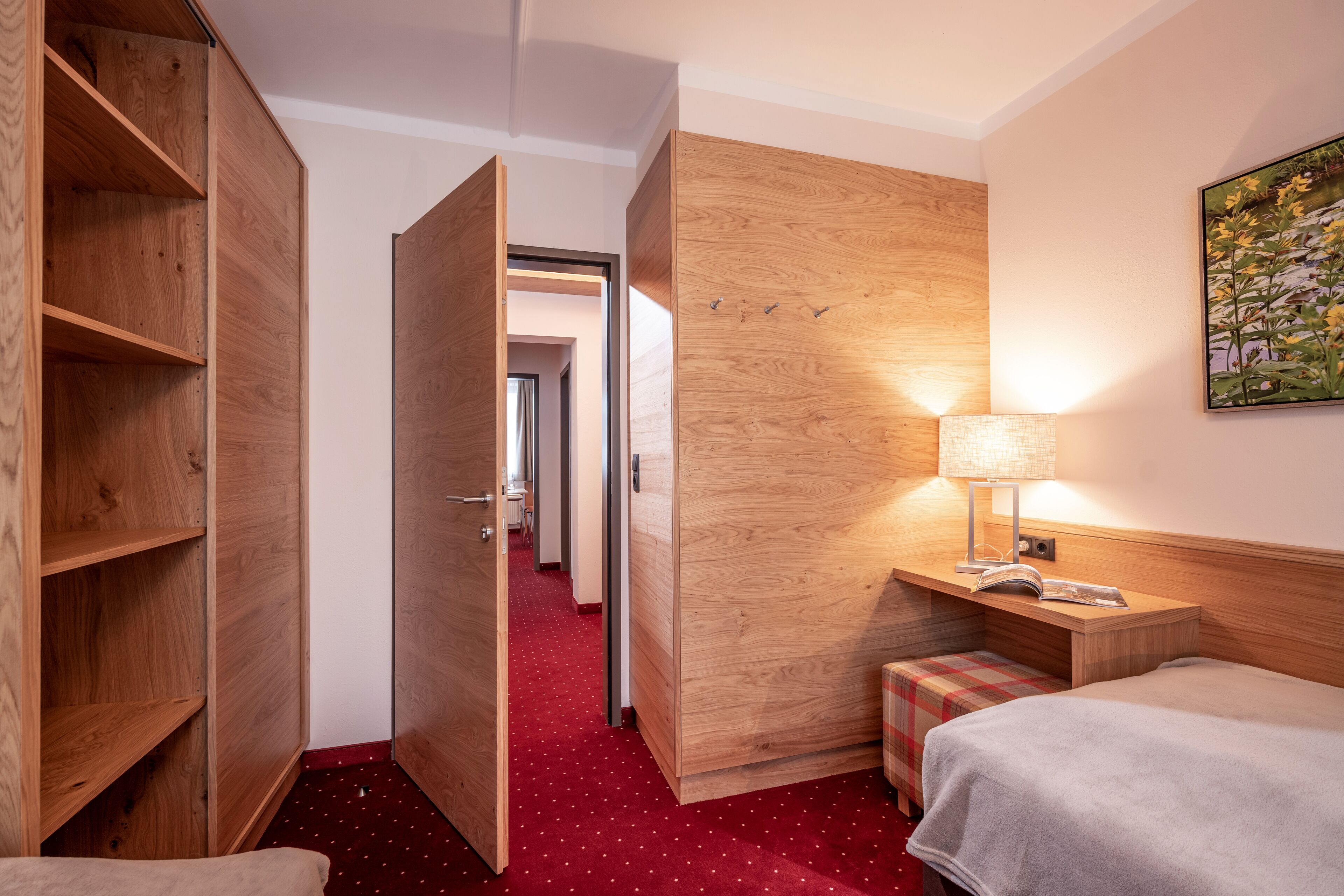Habitación de hotel con armario de madera, mesita de noche con lámpara, cama y alfombra roja.