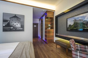 Habitación de hotel con cama, un banco, televisor y pasillo con un armario de madera y un cuadro enmarcado de un castillo.