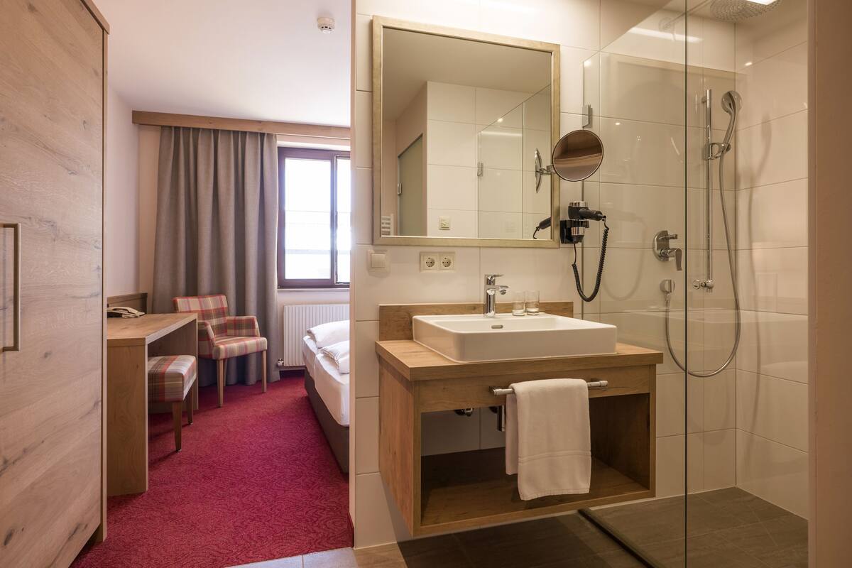 A hotel room with a wooden desk, a chair, a bed, a large mirror, a sink, and a shower.
