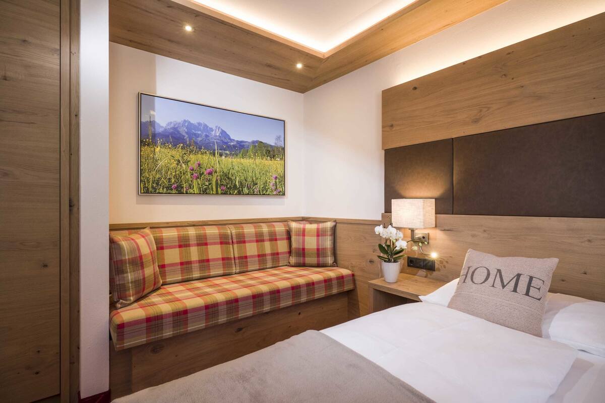 A bedroom with a bed, a plaid sofa, a nightstand with a lamp, a wall-mounted picture, and a wooden headboard.