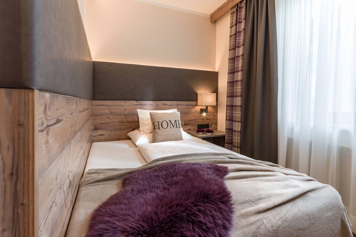 A modern bedroom with a wooden headboard, a bed with a 'HOME' pillow, a nightstand with a lamp, and sheer curtains.