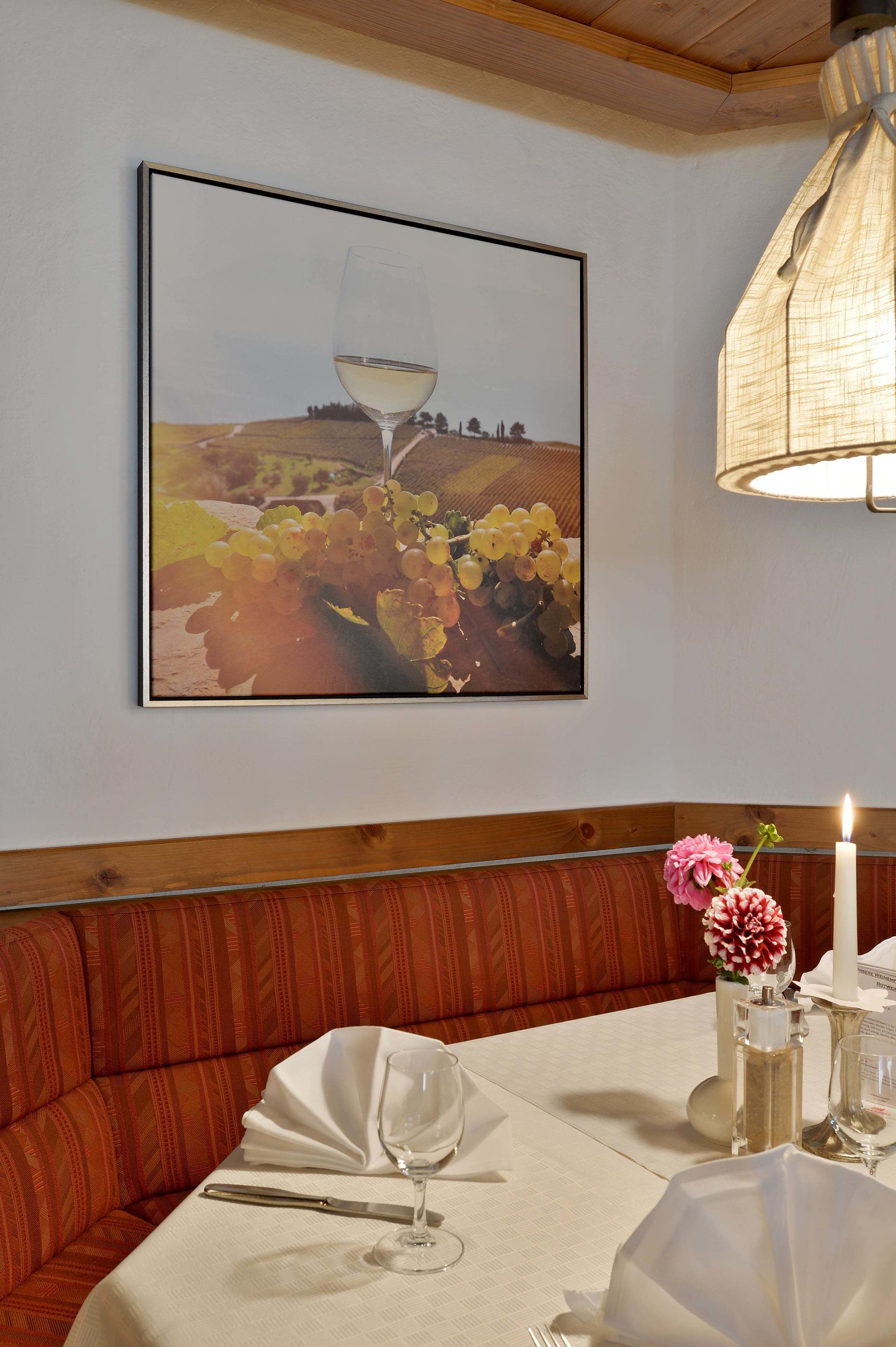 A dining area with a table set for two, a framed artwork of a wine glass and grapes, and a candle on the table.