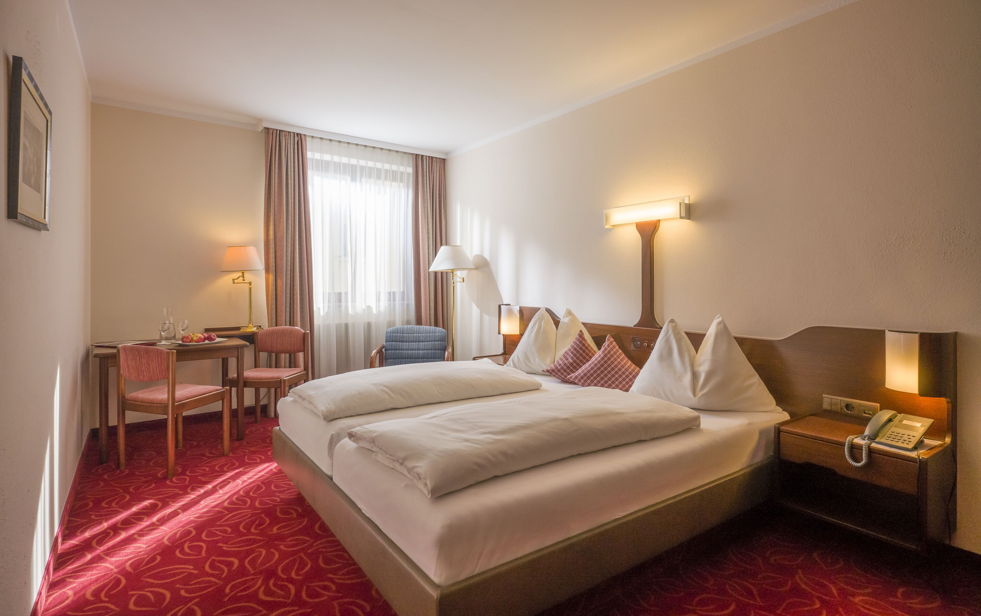 A hotel room with a large bed, a desk with a chair, a telephone, and a window with curtains.