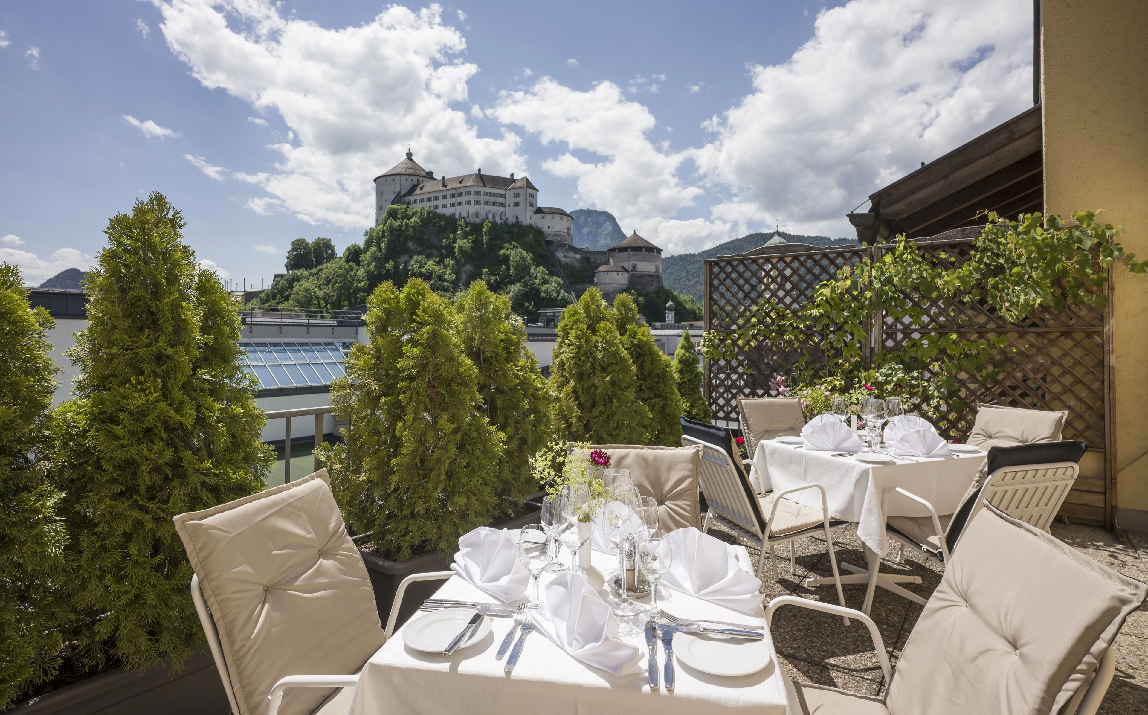 An outdoor dining area with tables set for two, overlooking a body of water and a historic castle.