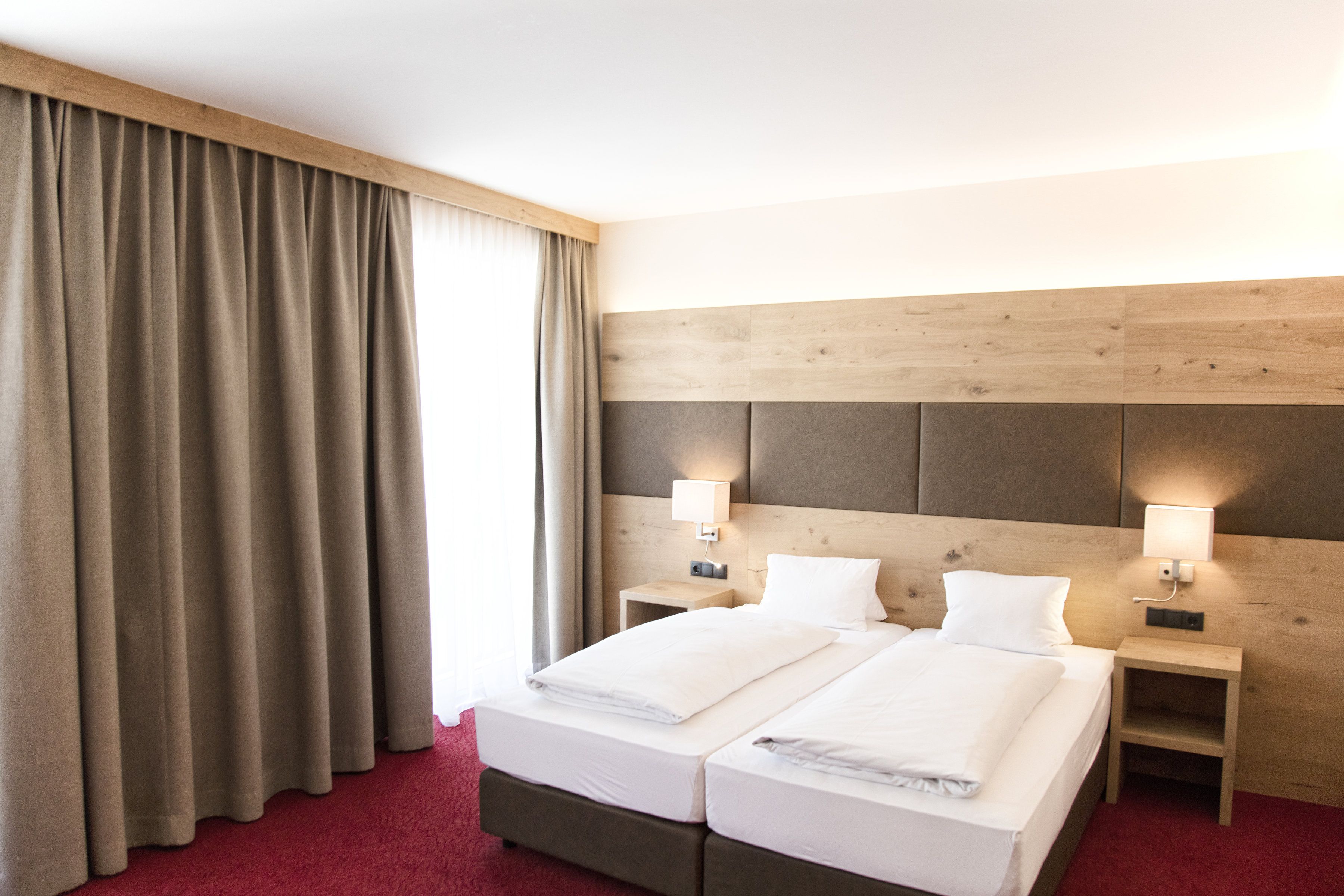 Una cama doble con cabecera de madera, dos mesitas de noche con lámparas, una alfombra roja y un ventanal con cortinas.