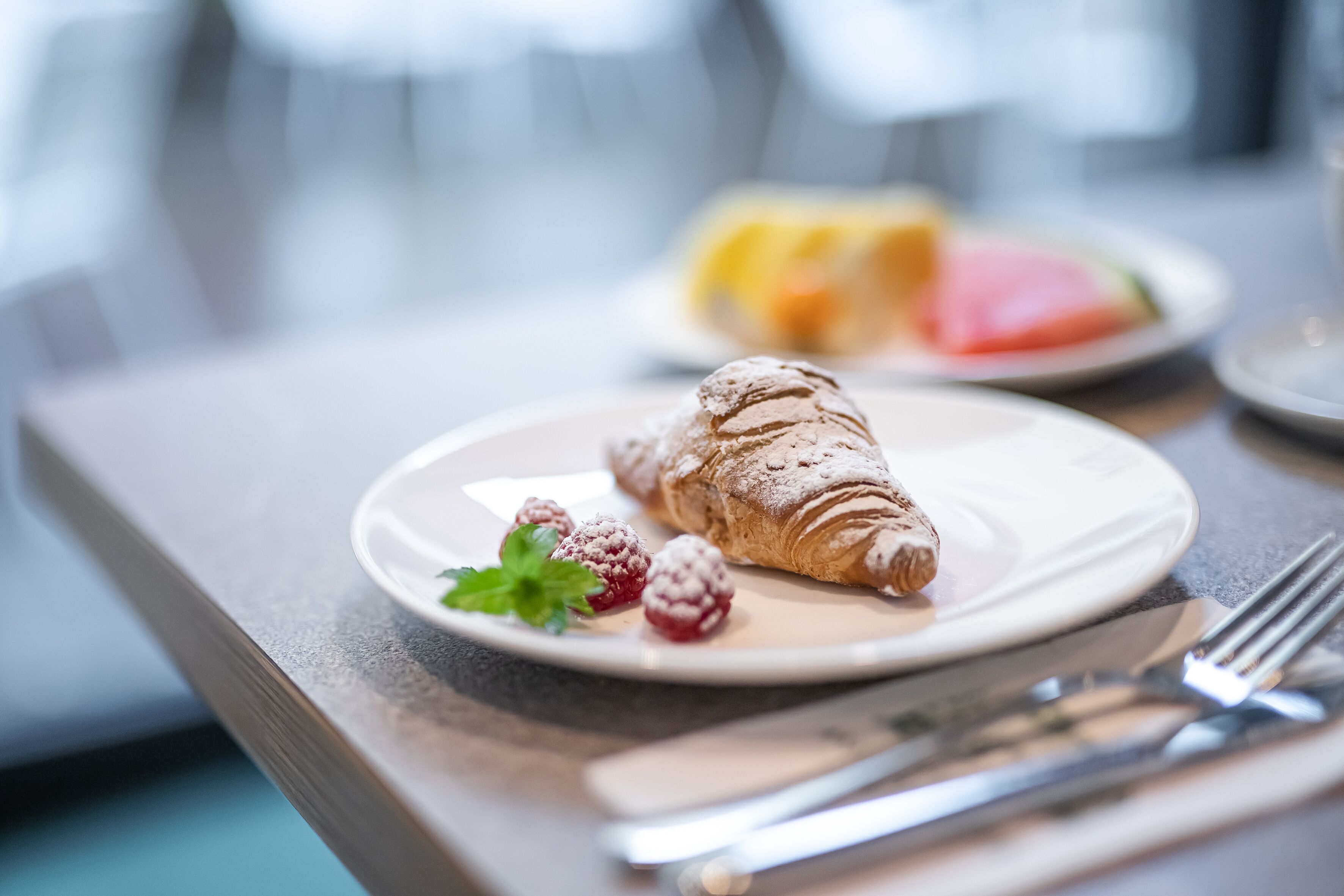 A croissant on a plate with raspberries and mint, a fork, and a knife on a table.