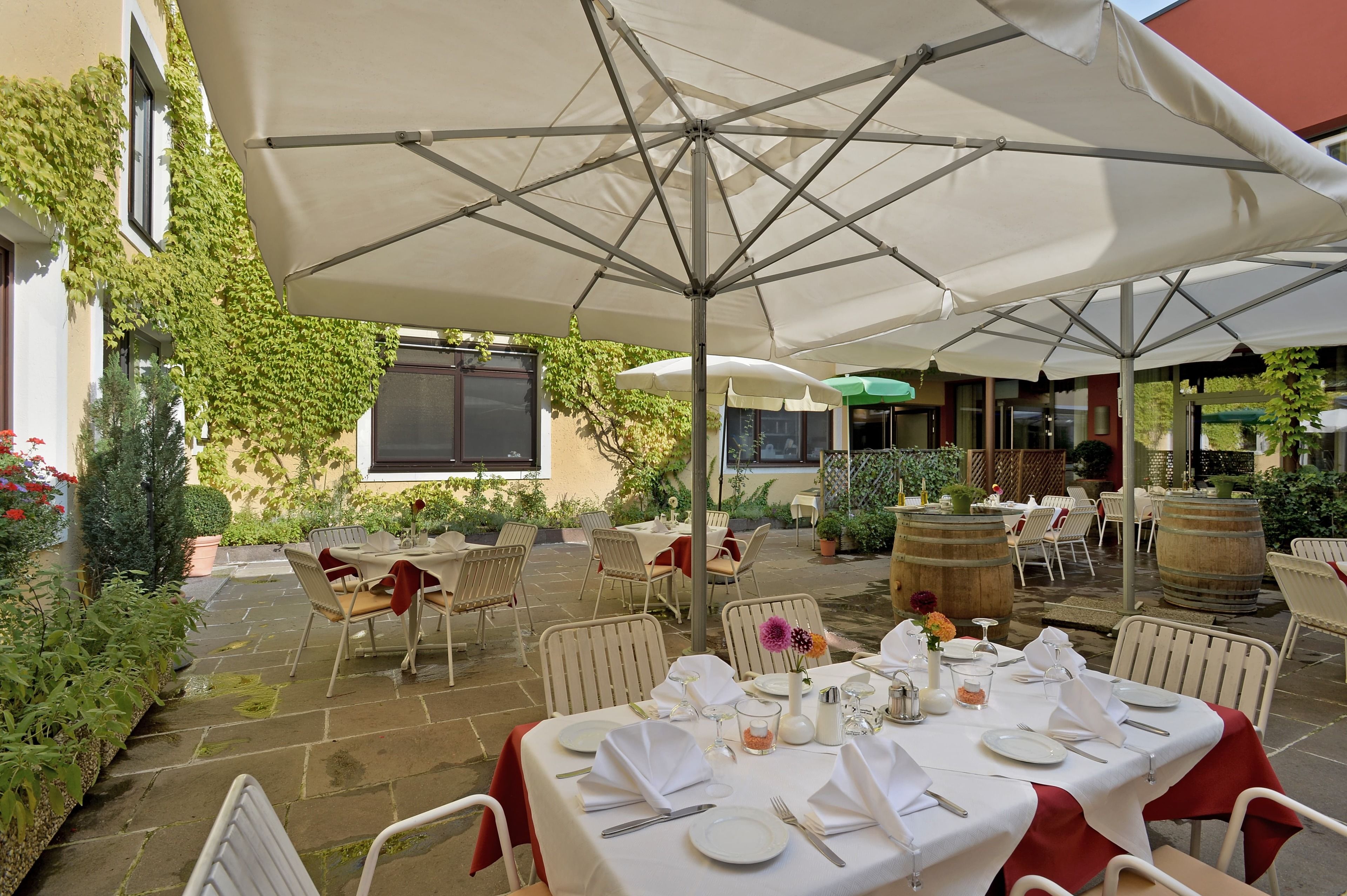 An outdoor dining area with tables set for a meal, surrounded by umbrellas and potted plants.