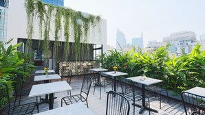 An outdoor patio with tables and chairs, surrounded by lush greenery and a modern building in the background. - Cherry Boutique Hotel (Ho Chi Minh City)