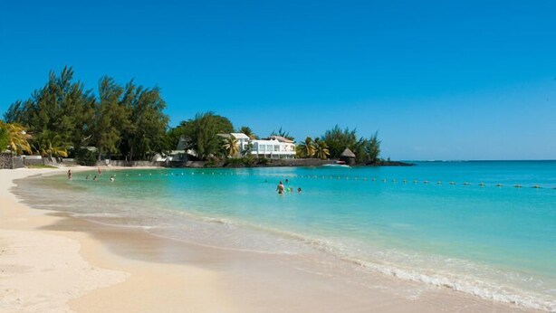 A beach with clear turquoise water, a sandy shore, and a building with a white facade. - Dodola Lodge (Grand-Baie)