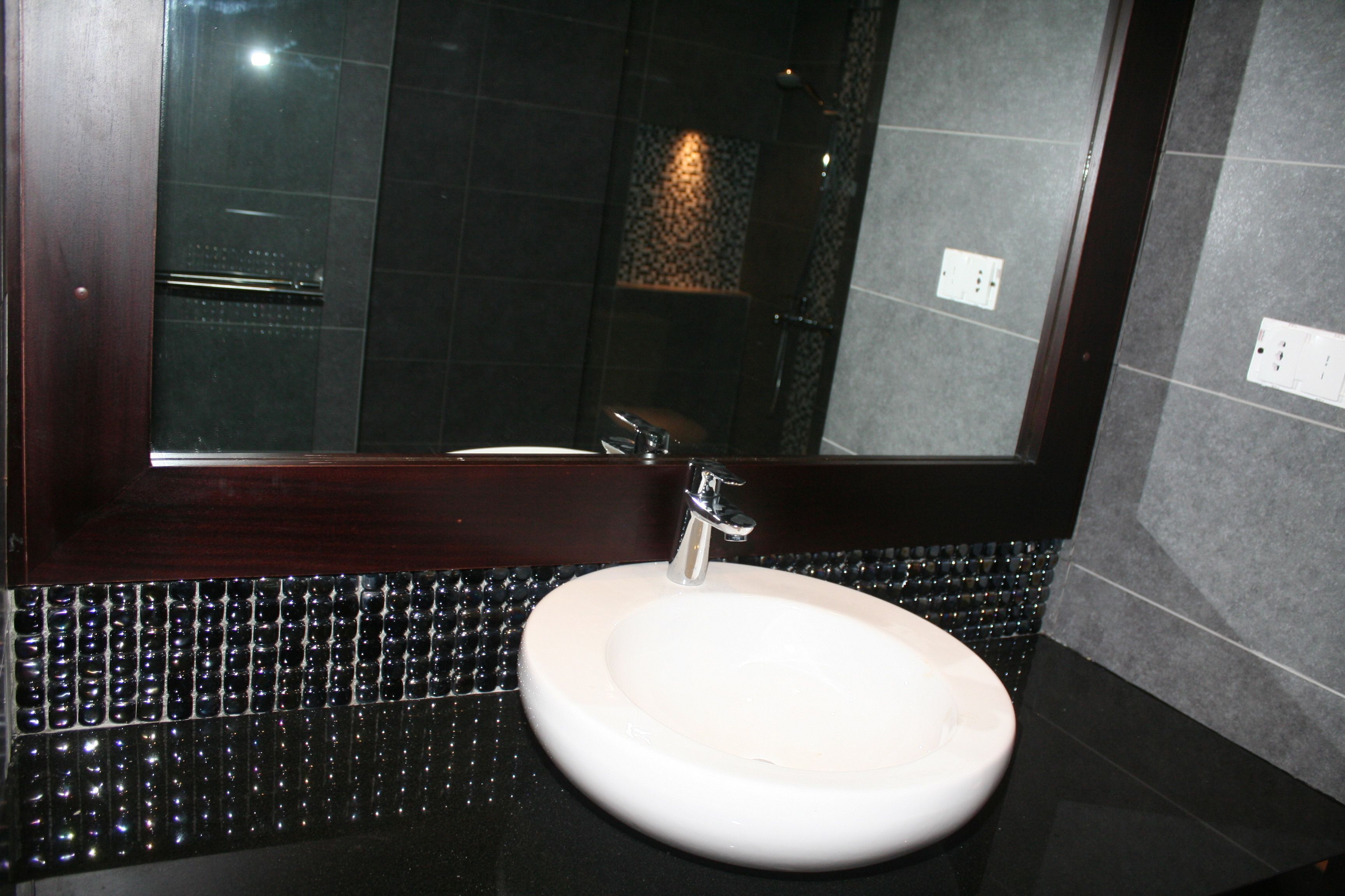 Un baño con un lavamanos redondo, paredes cubiertas de azulejos oscuros y un espejo grande.