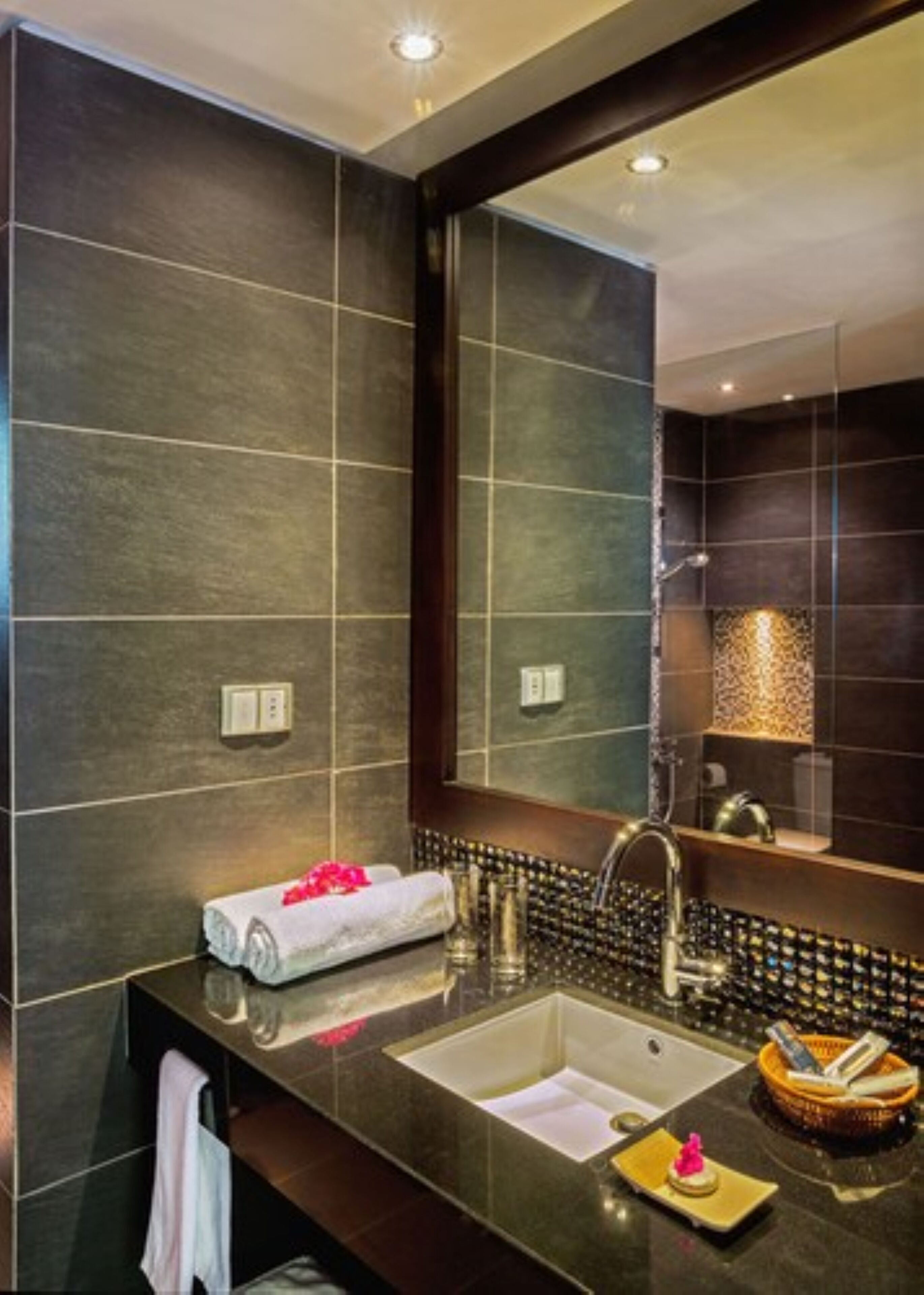 Un baño moderno con baldosas oscuras, un espejo grande, un lavamanos con una toalla y un estante con artículos de aseo.