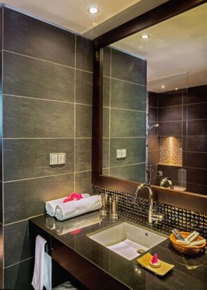 A modern bathroom with dark tiles, a large mirror, a sink with a towel, and a shelf with toiletries. - Dodola Lodge (Grand-Baie)