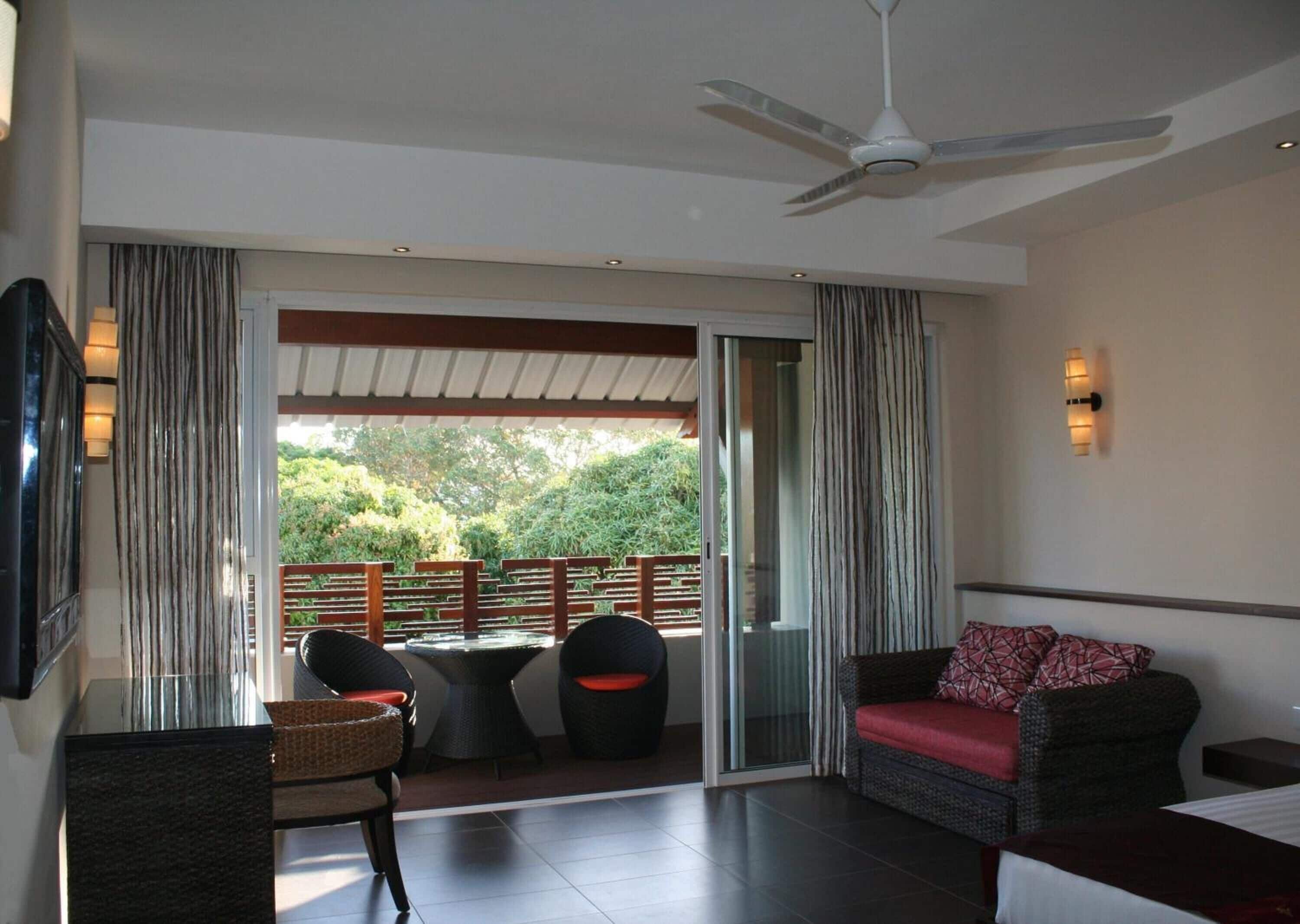 A modern hotel room with a balcony, a ceiling fan, a TV mounted on the wall, and a sofa with a red cushion.