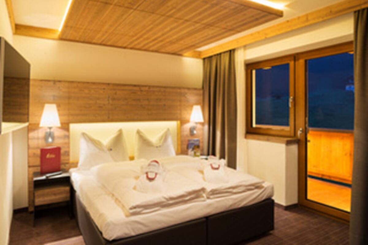 A hotel room with a wooden ceiling, a large bed with white linens, two bedside lamps, and a view of the outside through a large window.