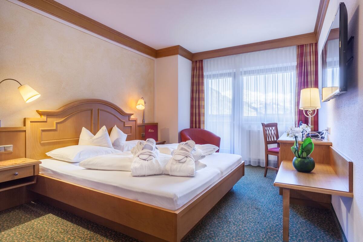 A hotel room with a neatly made bed, a desk with a lamp, a chair, and a small table with a vase of flowers.