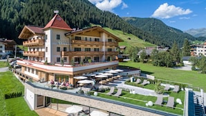 Un hotel con terraza, mobiliario exterior y montañas al fondo.