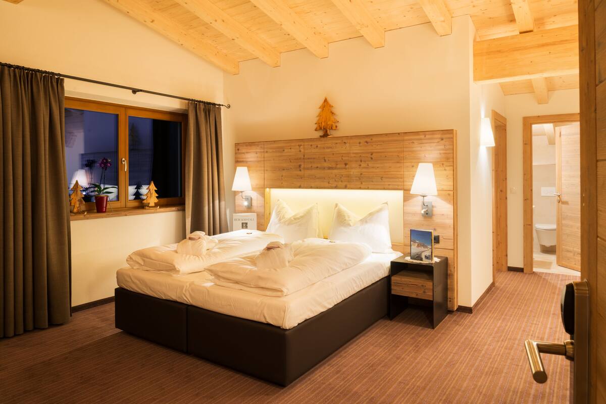 A hotel room with a wooden ceiling, a bed with white linens, two bedside tables with lamps, a window with curtains, and a small Christmas tree on the wall.