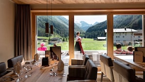 Un comedor con una mesa de madera preparada para una comida, ventanas amplias que ofrecen vistas a un paisaje montañoso, y una persona de pie junto a la ventana.