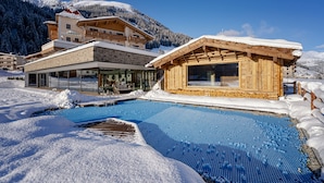 Zona de piscina exterior cubierta de nieve con cabañas de madera y montañas al fondo.