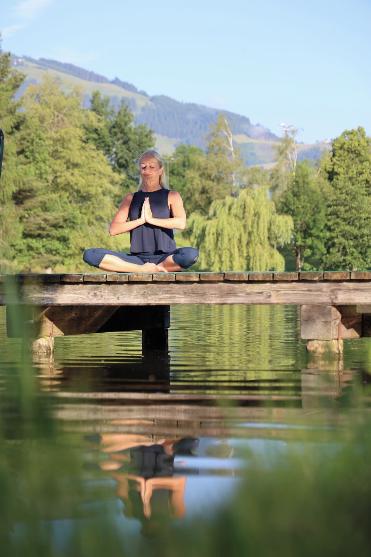 A person meditating on a wooden bridge over a calm body of water, surrounded by greenery and hills.