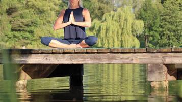 A person meditating on a wooden bridge over a calm body of water, surrounded by greenery and hills.